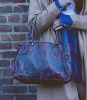 A person holding the Rockababy – a red leather everyday handbag with dual handles by Bed Stu, is wearing a tan coat and denim shirt while standing against a brick wall.