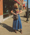 Woman in a casual outfit with a Rockababy graphic tee, denim skirt, and ankle boots, carrying a Bed Stu stylish practical bag, walking on a sunny sidewalk.