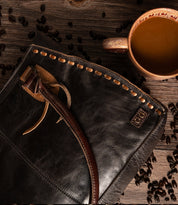 Rockababy compact vegetable-tanned leather bag with woven edge detail, next to a mug of coffee and scattered coffee beans on a wooden surface.