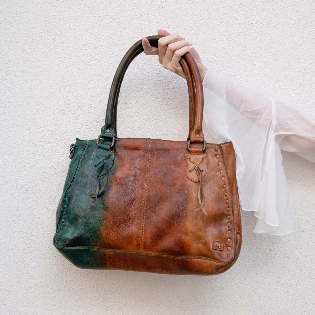 A hand holds the Bed Stu Rockababy bag, showcasing green and brown leather color blocking, stitched details, and a crossbody strap against a light textured wall.