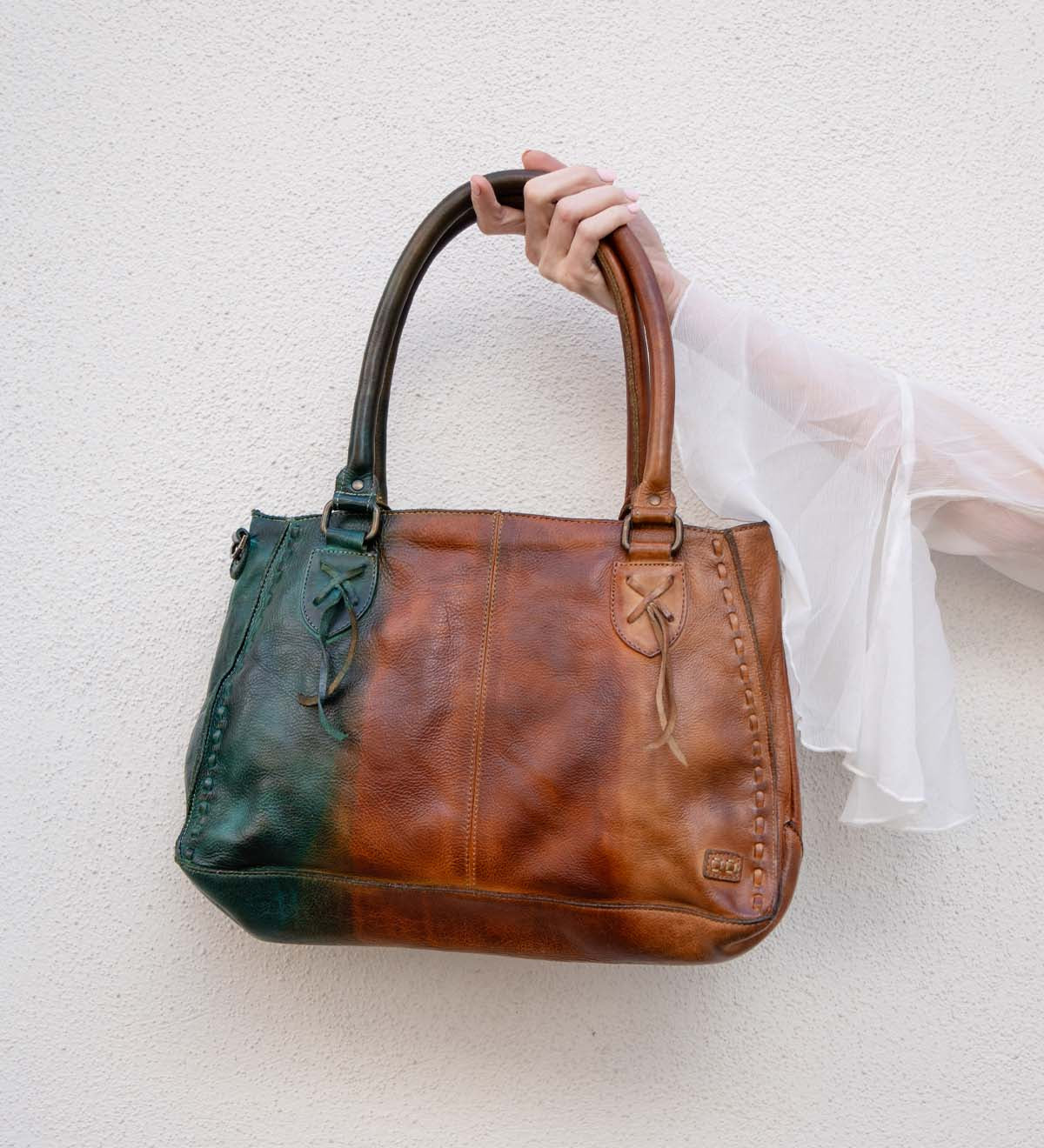 A hand holds the Bed Stu Rockababy bag, showcasing green and brown leather color blocking, stitched details, and a crossbody strap against a light textured wall.