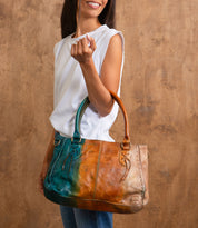 A woman stands against a wooden background, wearing a white sleeveless top and blue jeans, holding the Bed Stu Rockababy bag with padded shoulder straps and a blue-brown gradient design.