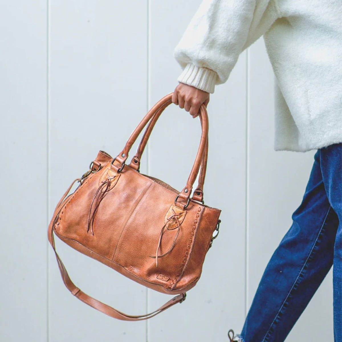 A person in a white sweater and blue jeans holds the Bed Stu Rockababy bag, featuring two handles, a crossbody strap, and a compact everyday design, against a light background.
