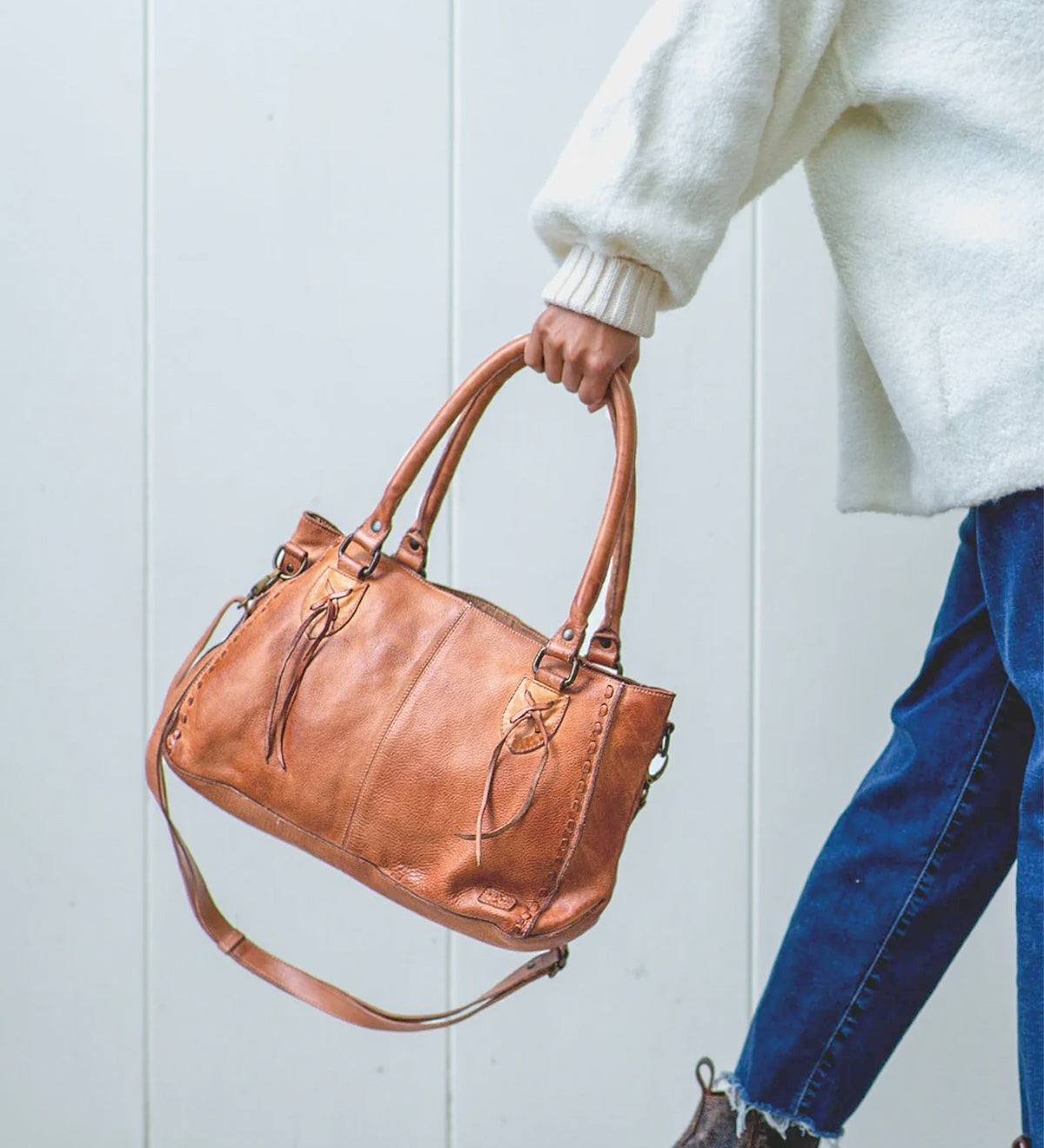 A person in a white sweater and blue jeans holds the Bed Stu Rockababy bag, featuring two handles, a crossbody strap, and a compact everyday design, against a light background.