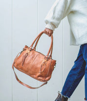 A person in a white sweater and blue jeans holds the Bed Stu Rockababy bag, featuring two handles, a crossbody strap, and a compact everyday design, against a light background.