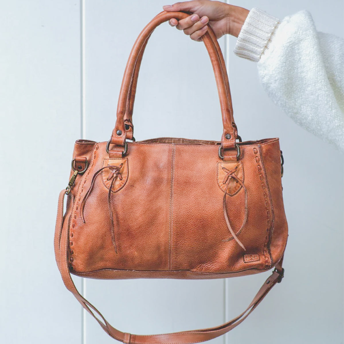 A hand holds a brown leather Bed Stu Rockababy bag with dual handles, crossbody strap, and decorative stitching against a plain light background.