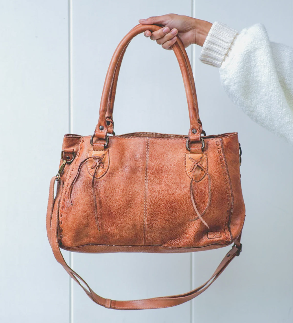 A hand holds the Rockababy by Bed|Stü—a compact leather bag crafted from vegetable-tanned leather, with dual handles, decorative stitching, and an attached crossbody strap—set against a plain white background.