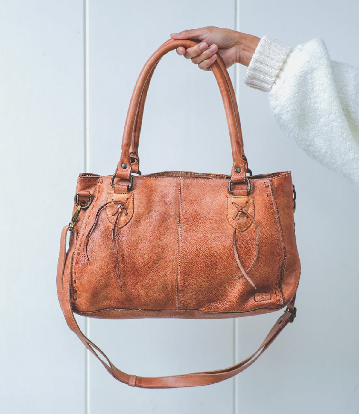 A hand holds a brown leather Bed Stu Rockababy bag with dual handles, crossbody strap, and decorative stitching against a plain light background.