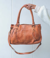 A hand holds a brown leather Bed Stu Rockababy bag with dual handles, crossbody strap, and decorative stitching against a plain light background.