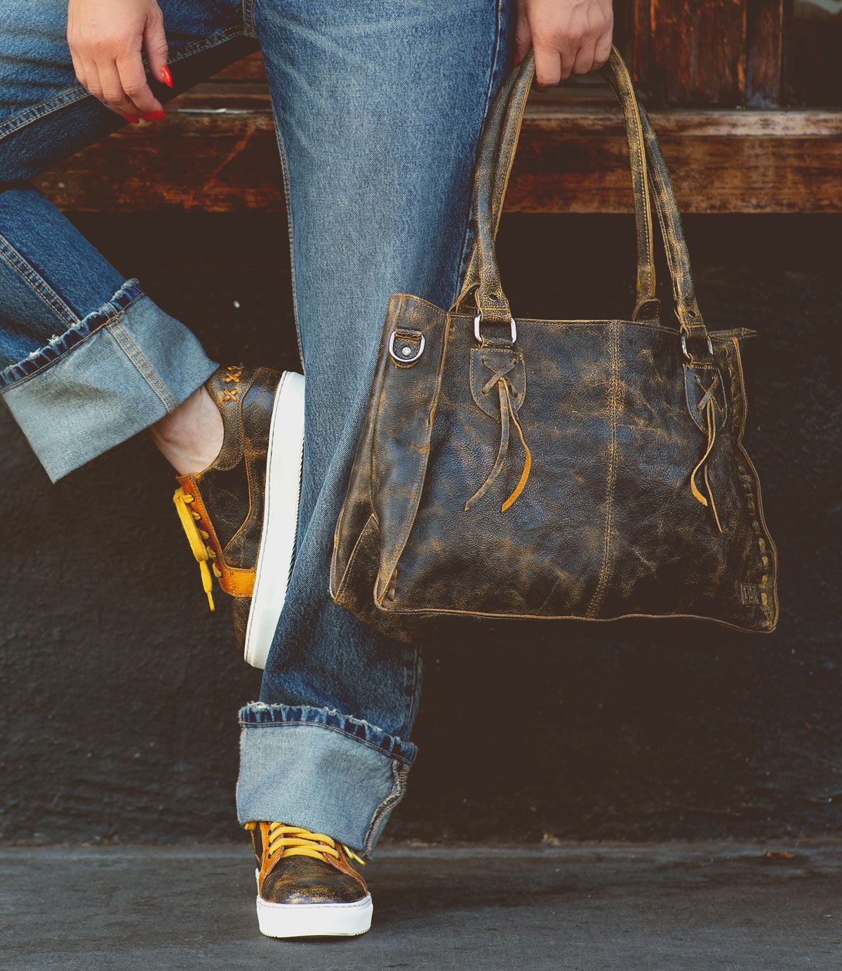 Person wearing blue jeans and sneakers holds a Bed Stu Rockababy everyday bag while leaning against a wooden surface.