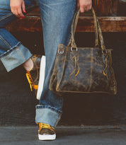 Person wearing blue jeans and sneakers holds a Bed Stu Rockababy everyday bag while leaning against a wooden surface.