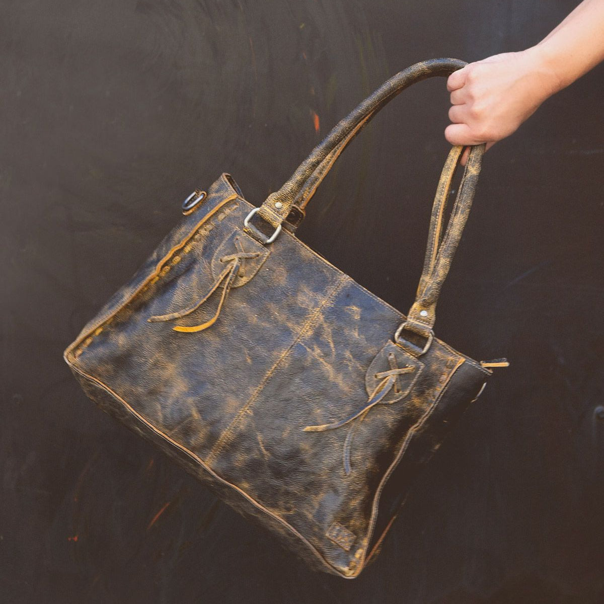 A hand holding the Bed Stu Rockababy distressed brown leather tote bag with two handles and decorative tassels against a dark background, perfect for an everyday bag.