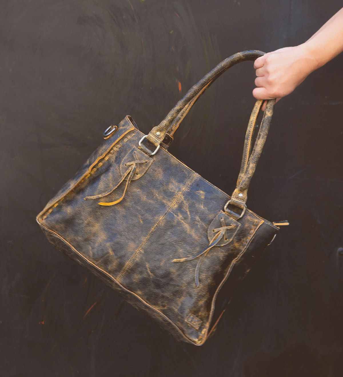 A hand holding the Bed Stu Rockababy distressed brown leather tote bag with two handles and decorative tassels against a dark background, perfect for an everyday bag.