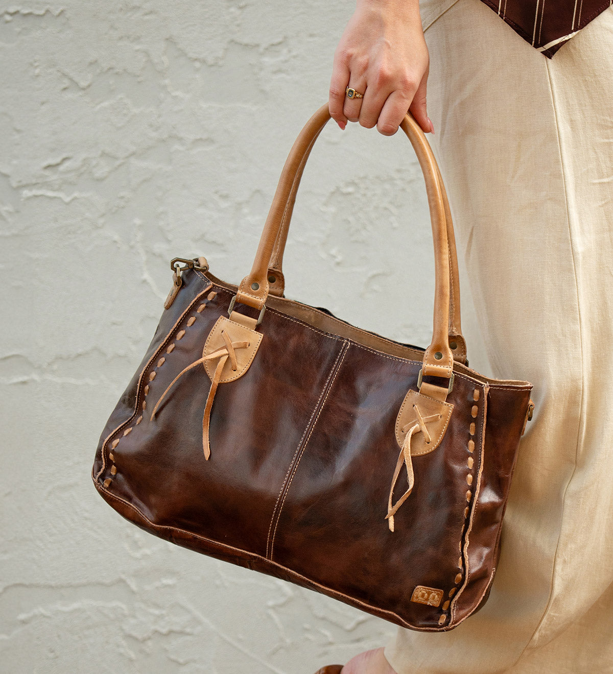 A person in beige pants holds the Bed|Stü Rockababy, a compact leather bag crafted from vegetable-tanned leather, featuring tan handles and decorative stitching, against a light textured wall.