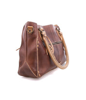 The Bed Stu Rockababy is a compact brown leather handbag with tan handles, visible stitching, and a side zipper pocket. Includes an adjustable crossbody strap—ideal for everyday essentials. Shown on a white background.