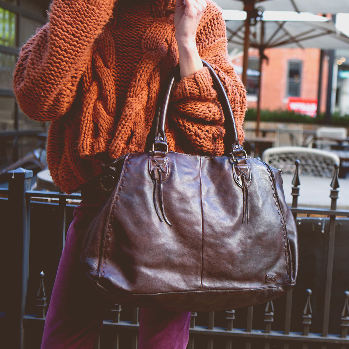 A person in an orange sweater holds stylish sunglasses and the popular large brown leather Rockaway handbag from Bed Stu, featuring an optional shoulder strap, outdoors.