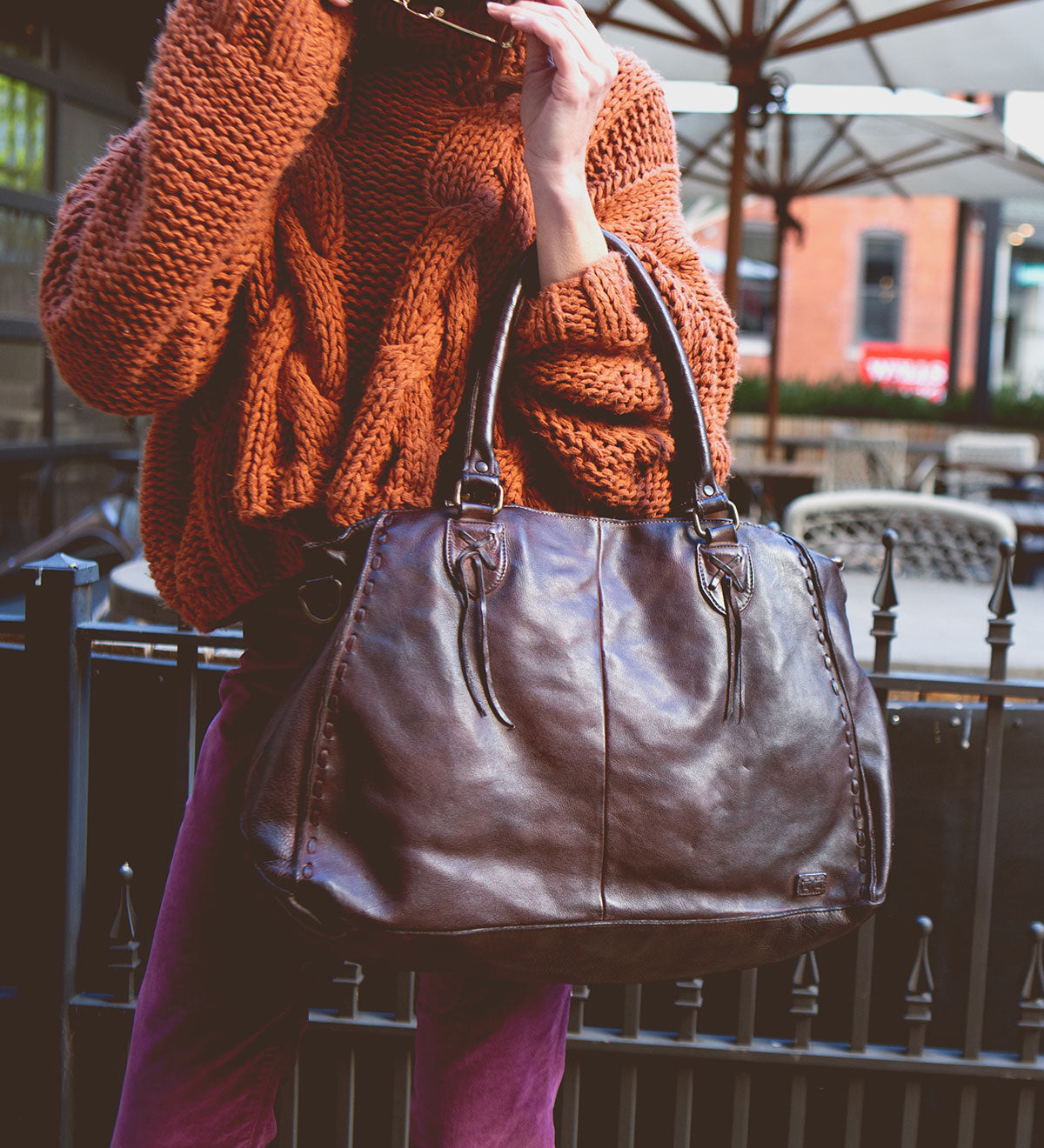 A person in an orange sweater holds stylish sunglasses and the popular large brown leather Rockaway handbag from Bed Stu, featuring an optional shoulder strap, outdoors.