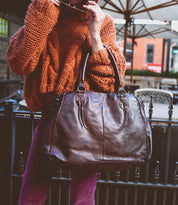 A person in an orange sweater holds stylish sunglasses and the popular large brown leather Rockaway handbag from Bed Stu, featuring an optional shoulder strap, outdoors.