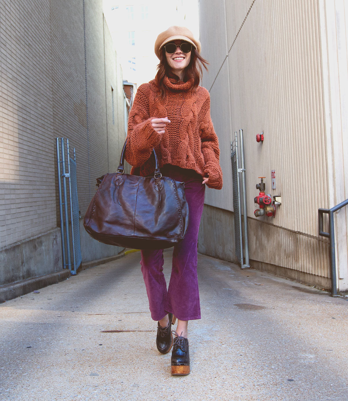 A person strolls through an alley wearing a beige hat, sunglasses, an orange sweater, purple pants, and platform shoes. They're carrying their best-selling Rockaway bag from Bed Stu with an optional shoulder strap and a convenient zip-pocket divider.