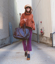 A person strolls through an alley wearing a beige hat, sunglasses, an orange sweater, purple pants, and platform shoes. They're carrying their best-selling Rockaway bag from Bed Stu with an optional shoulder strap and a convenient zip-pocket divider.