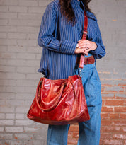 A person in a blue striped shirt and denim pants holds the Bed Stu Rockaway bag, featuring a zip-pocket divider and shoulder strap, standing in front of a weathered brick wall.