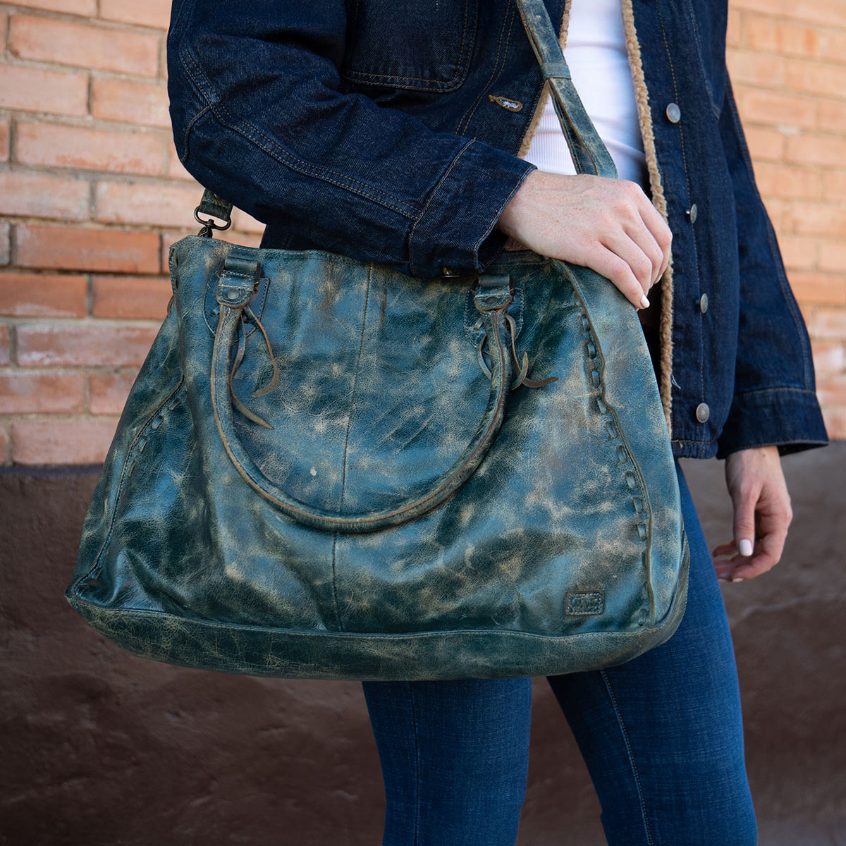 A person in a dark denim jacket and jeans stands against a brick wall, holding Bed Stu’s best-selling Rockaway—a large distressed green leather handbag with shoulder straps and a zip-pocket divider.