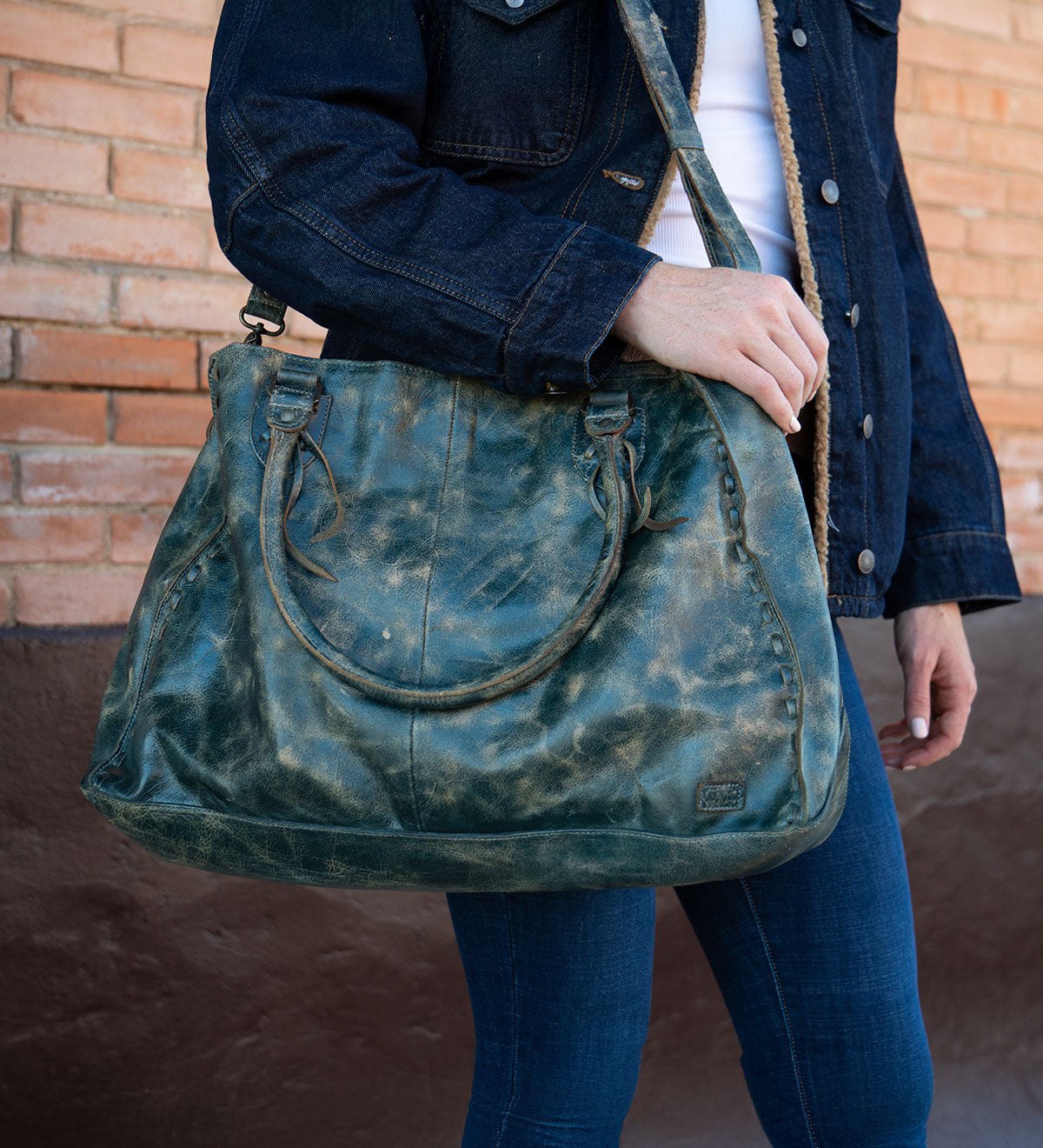 A person in a dark denim jacket and jeans stands against a brick wall, holding Bed Stu’s best-selling Rockaway—a large distressed green leather handbag with shoulder straps and a zip-pocket divider.