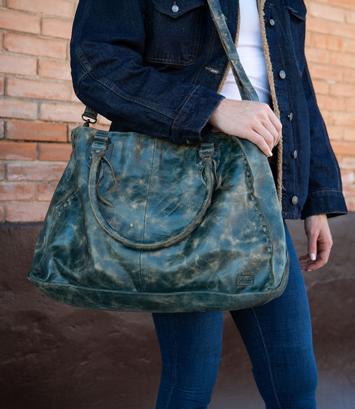 A person in a dark denim jacket and jeans stands against a brick wall, holding Bed Stu’s best-selling Rockaway—a large distressed green leather handbag with shoulder straps and a zip-pocket divider.
