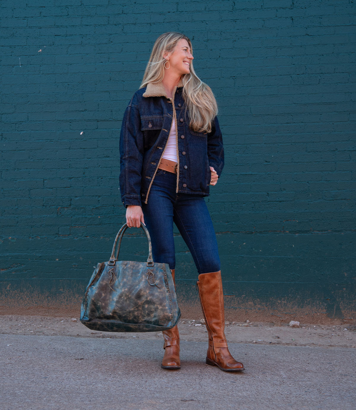 A woman stands in front of a teal brick wall, wearing a denim jacket, jeans, brown boots, and holding the Bed Stu Rockaway bag with a practical shoulder strap.