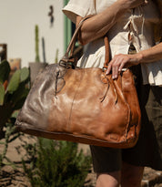 A person holding the Bed Stu Rockaway, a large brown full-grain leather handbag with two handles, stands outdoors near cacti and greenery.