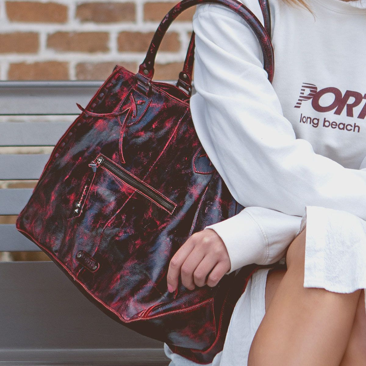 Person sitting on a bench holding a best-selling Rockaway handbag in a black and red pattern with a small front zipper compartment, wearing a white long-sleeve shirt. A brick wall is visible in the background, exemplifying the perfect blend of style and functionality characteristic of Bed Stu designs.