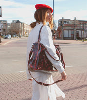 A woman in a white coat and red beret stands on a city street, holding her Rockaway bag by Bed Stu on her shoulder, with buildings and a bar visible in the background.