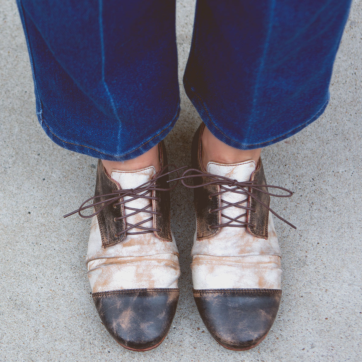 A person wearing blue jeans and vintage-style lace-up shoes with a worn appearance, standing on a concrete surface, exudes Rumba II by Bed Stu flair.