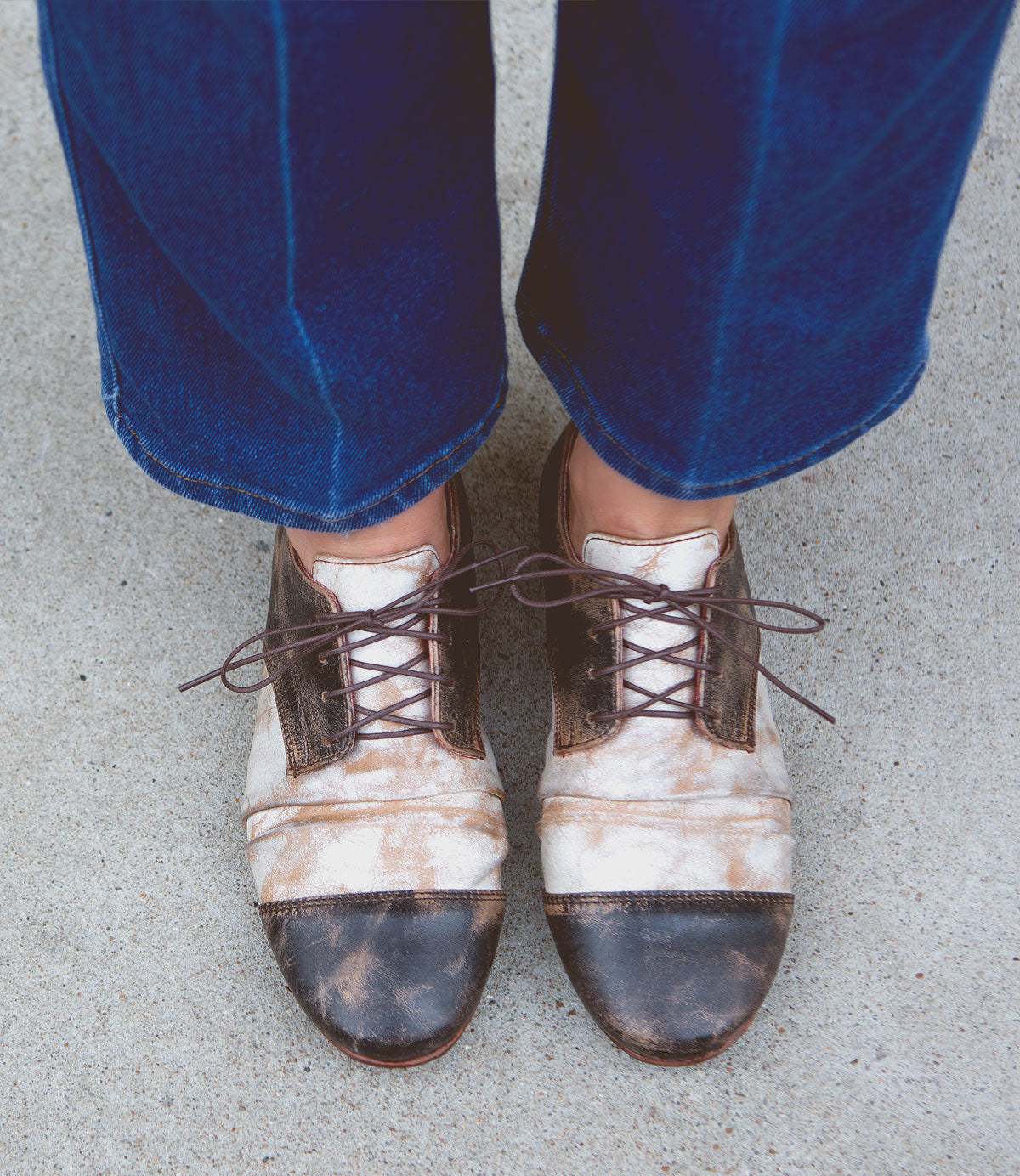 A person wearing blue jeans and vintage-style lace-up shoes with a worn appearance, standing on a concrete surface, exudes Rumba II by Bed Stu flair.