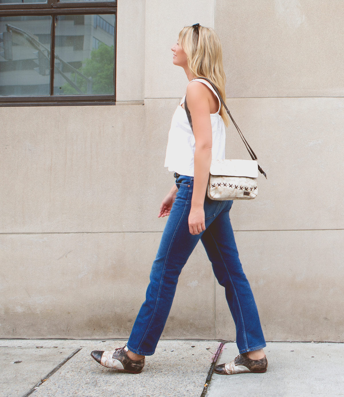 A person in blue jeans, a white top, and sandals walks on a sidewalk past a building with a window. They carry a Rumba II by Bed Stu, featuring buttery soft leather that adds an element of luxury to their casual ensemble.