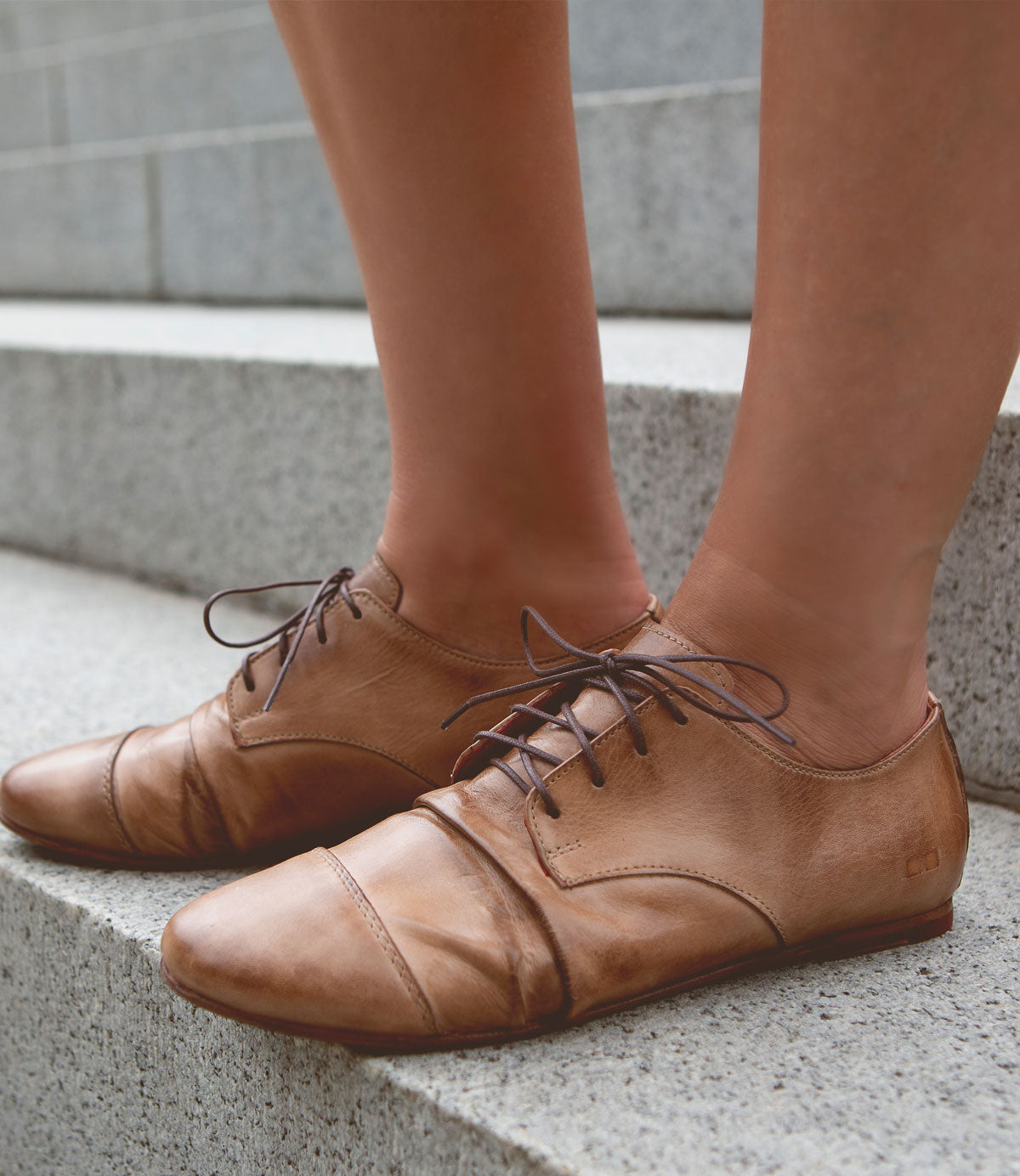 A person wearing brown lace-up leather shoes stands on a set of concrete steps, exuding a classic touch with their Bed Stu Rumba II ensemble.