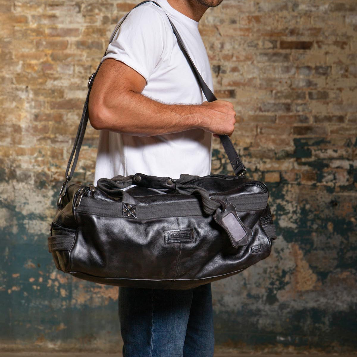 A man in a white t-shirt and jeans stands against a worn brick wall, carrying the Bed Stu Ruslan, a large black leather duffle, over his shoulder.