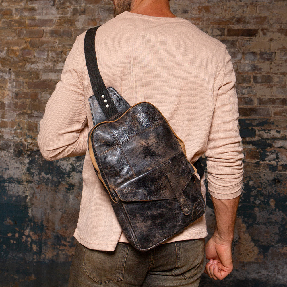 Person wearing a dark leather sling bag with a wide shoulder strap over a light textured shirt.