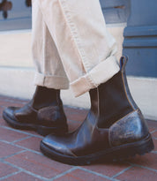 A person wearing the Bed Stu Salatiel, exuding timeless style with their brown leather finish and unmatched comfort, paired with rolled-up beige pants on a brick sidewalk.