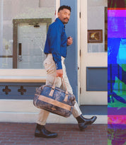 A man in a blue shirt and beige pants is walking on a brick sidewalk, carrying a brown leather bag, his stride confident in the Salatiel Chelsea boots by Bed Stu with a textured sole.