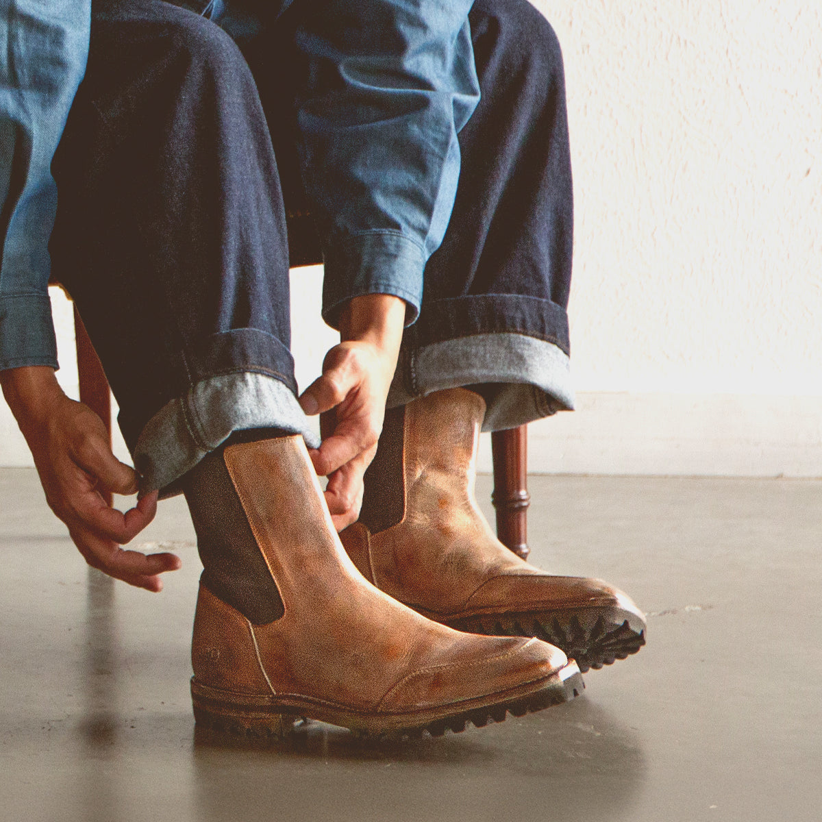 A person wearing Bed Stu Salatiel brown Chelsea boots with a textured sole and rolled-up jeans sits on a chair, adjusting their footwear.