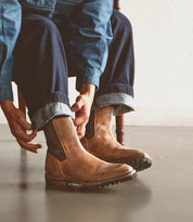 A person wearing Bed Stu Salatiel brown Chelsea boots with a textured sole and rolled-up jeans sits on a chair, adjusting their footwear.