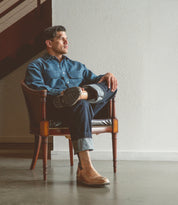 A person sits in a chair, wearing a denim shirt and jeans, looking sideways with crossed legs. A wooden staircase contrasts the white textured wall. Their Barton leather high-top sneakers by Bed Stu feature breathable insoles for perfect comfort and style integration.