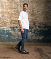 A man stands in front of a weathered brick wall, looking slightly to the left, wearing Bed Stu Samosa elegant men's shoes with a white t-shirt and blue jeans.