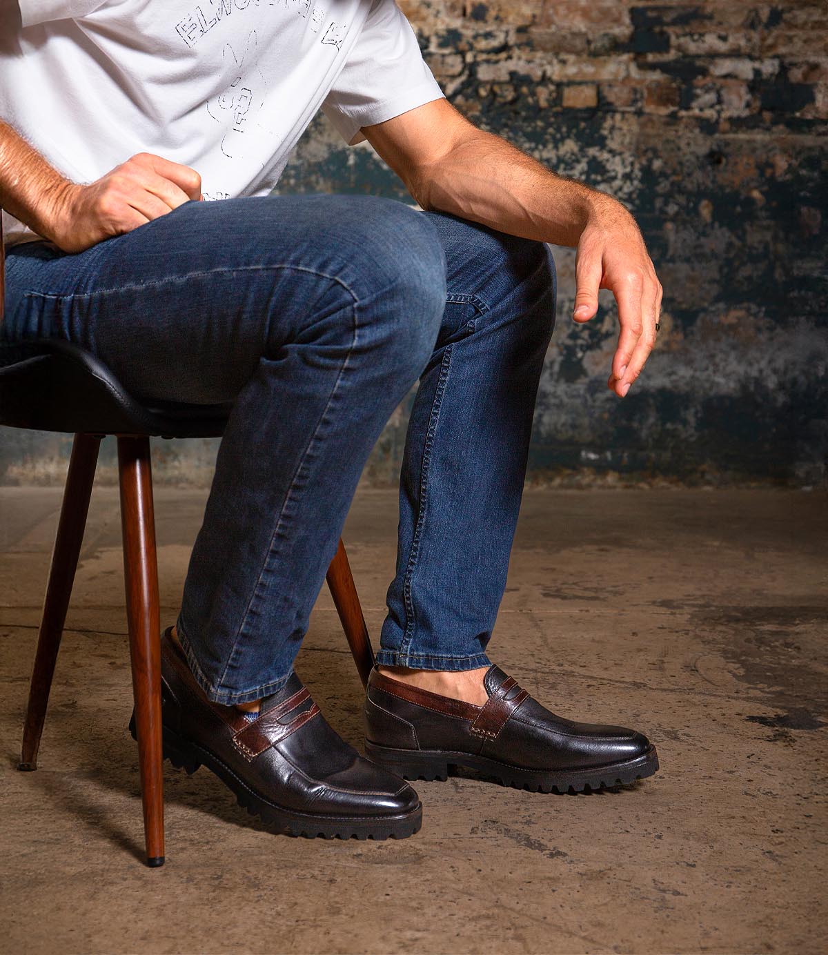 A man in a white t-shirt and blue jeans sits on a chair wearing Bed Stu’s Samosa—a pair of dark brown leather loafers with textured soles—set against a rustic background.