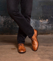 A person stands with legs crossed on a concrete floor against a textured wall, wearing dark jeans and Bed Stu's Sandro II tan handcrafted leather wingtip shoes.