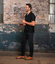 A man in a short-sleeve black shirt, black pants, and Bed Stu Sandro II handcrafted leather shoes stands smiling against a worn industrial brick wall.