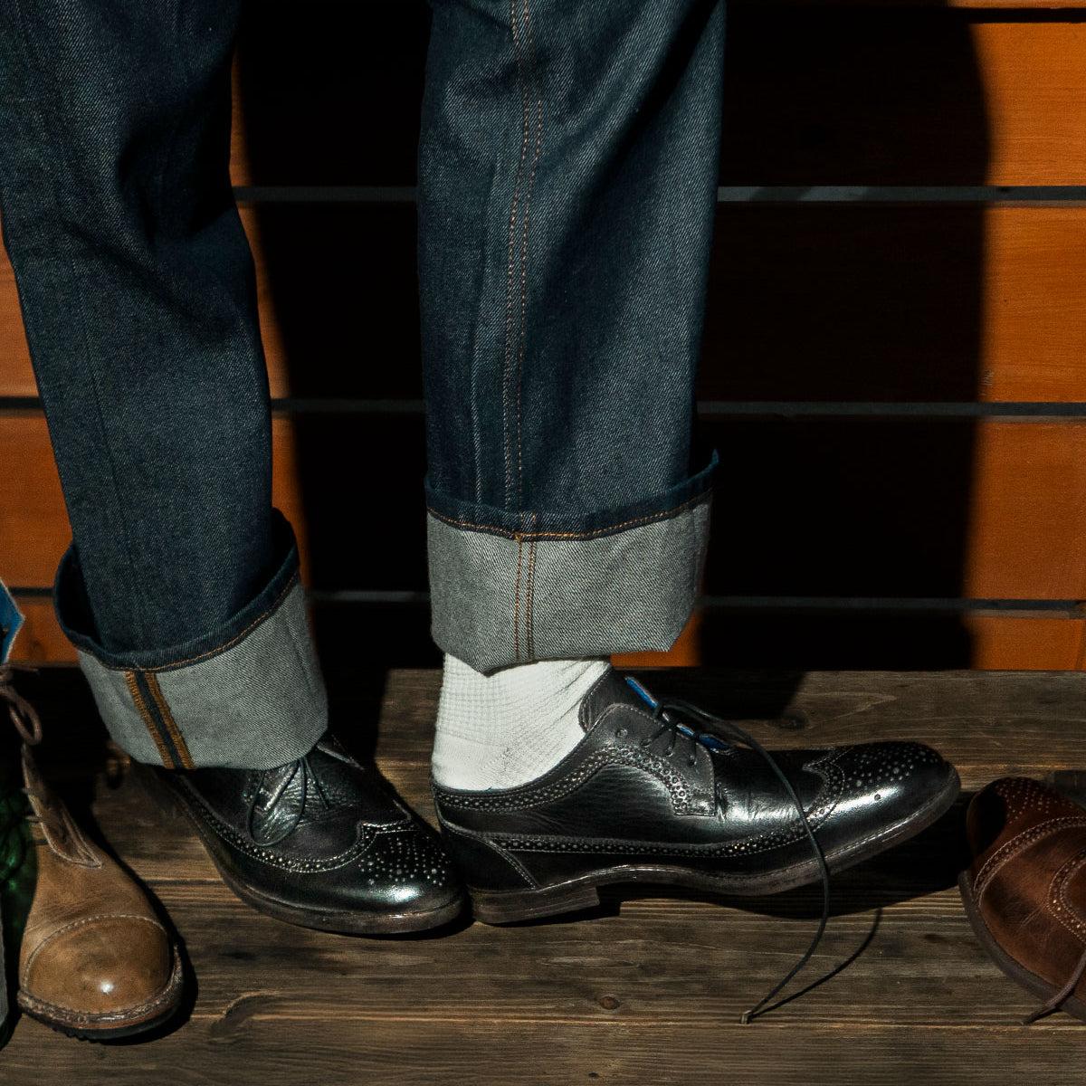 A person wearing cuffed jeans and black brogue shoes with white socks stands on a wooden floor, showcasing the intricate brogue details. Nearby, the Sandro II, a pair of handcrafted leather men's office shoes with a wingtip toe from Bed Stu, rests invitingly.