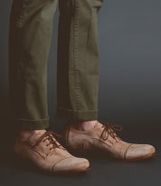 Person wearing olive green pants and light brown Bed Stu Sandstorm dress shoes with laces, elegantly crafted with a suede upper, standing against a dark background.