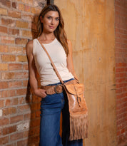 A woman in a sleeveless white top and blue jeans stands against a brick wall, wearing the Bed Stu Sandy Lane II—a handmade fringe crossbody bag crafted from vegetable-tanned leather—paired with a classic brown belt.
